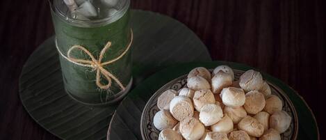 Un verre avec une paille et des glaçons, attaché avec une ficelle, posé sur une feuille verte. À côté, on aperçoit un bol rempli de biscuits ronds et blancs, disposés sur une assiette décorative.