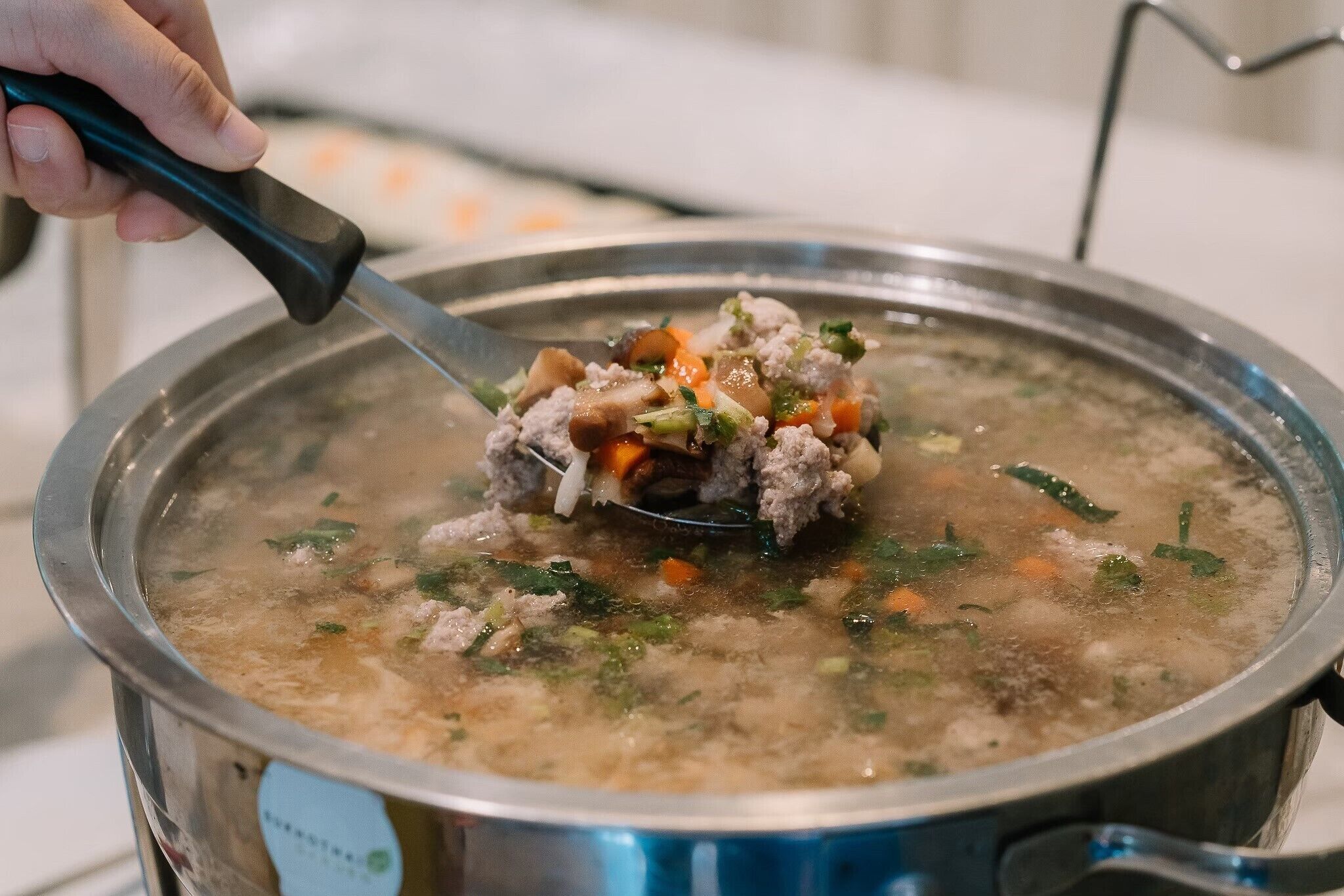 Uma pessoa mexendo uma panela de sopa com ingredientes visíveis como carne, cenouras e ervas.