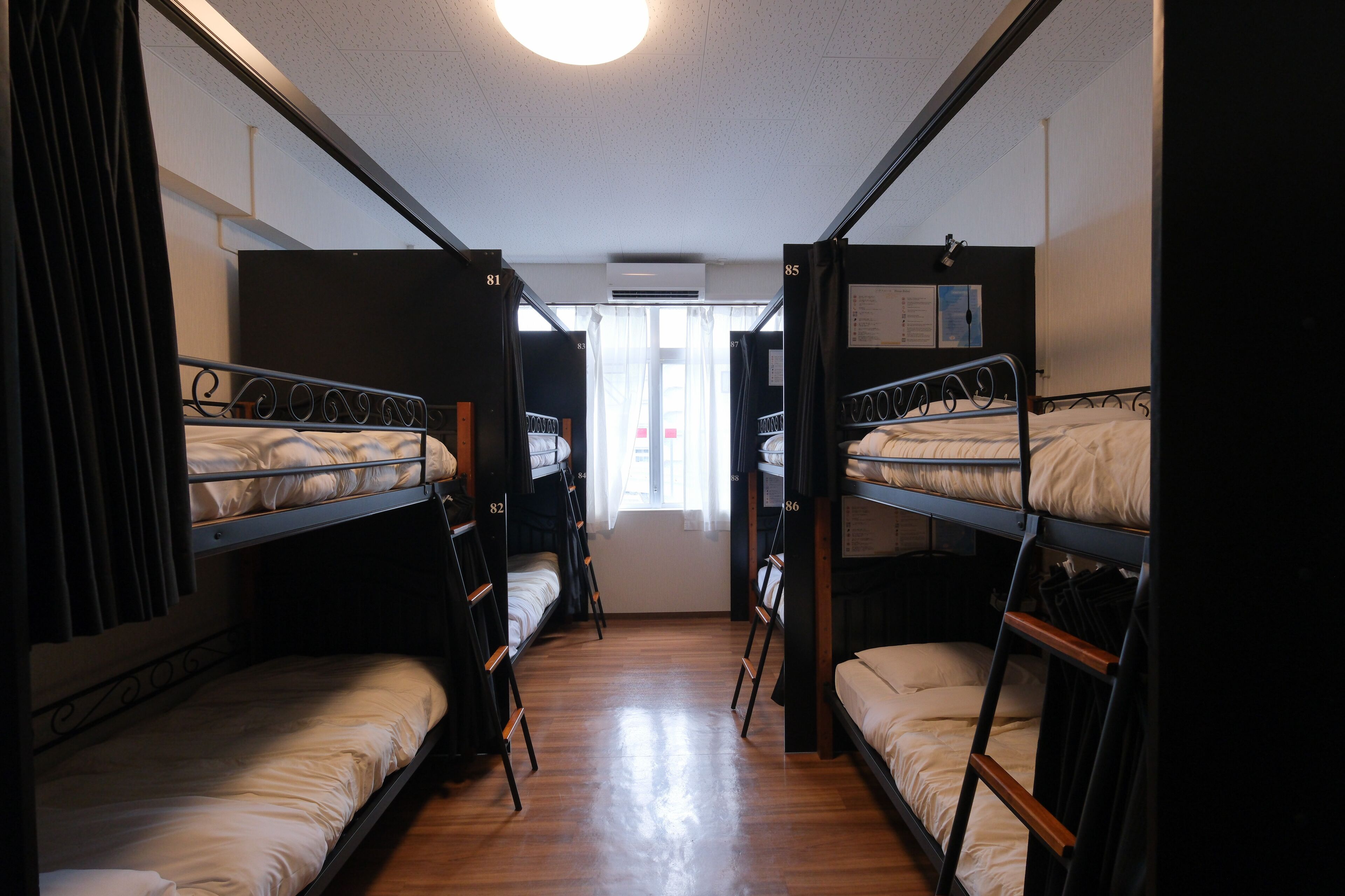 Un pasillo estrecho con literas a ambos lados, numeradas del 81 al 86, que conduce a una ventana con cortinas.