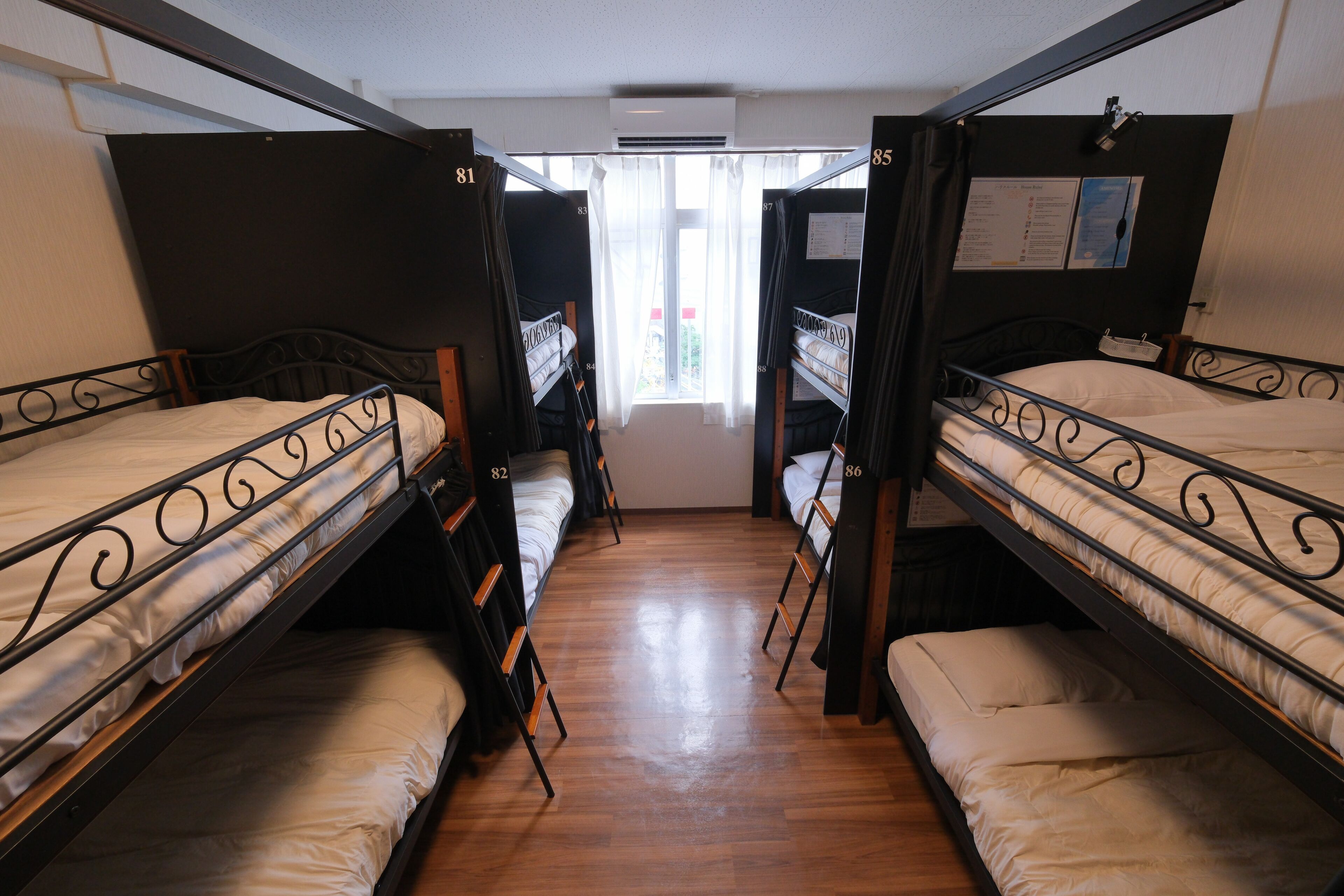 A dormitory room with bunk beds numbered 81, 82, 83, 84, and 85, wooden floors, and a window with curtains.