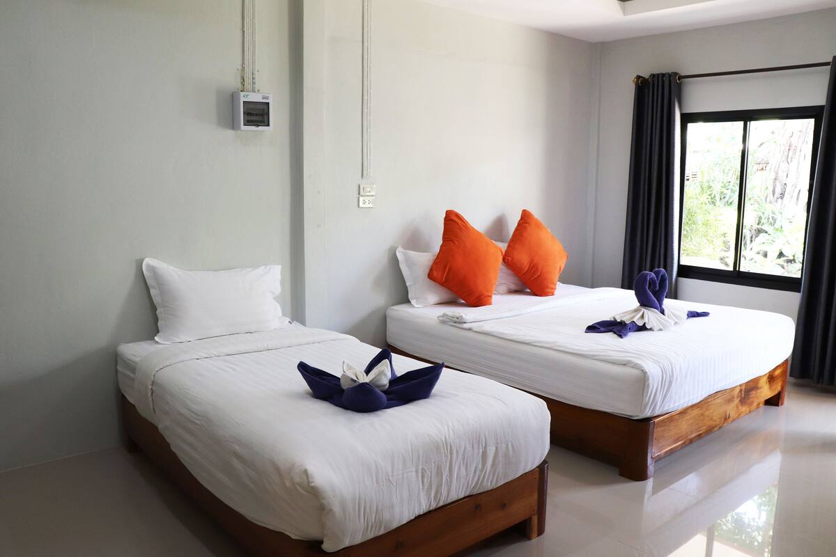 Dos camas individuales con ropa de cama blanca y almohadas naranjas, un cabecero de madera, un equipo de aire acondicionado pequeño montado en la pared y una ventana con cortinas oscuras.