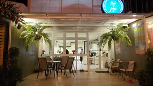 A hostel with a covered outdoor seating area, potted plants, and a neon sign indicating it is a 'Hub of Joys'. - Hub of Joys Hostel (Ko Lanta)