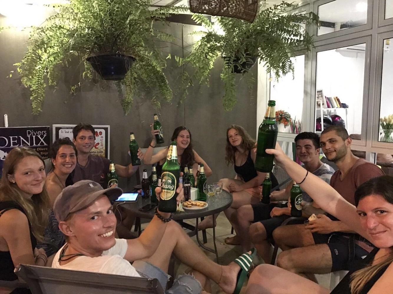 A group of people sitting around a table with bottles of Chang beer, in a room with a large window and hanging plants.