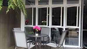 A covered patio area with a table and chairs, large windows, and a hanging plant. - Hub of Joys Hostel (Ko Lanta)