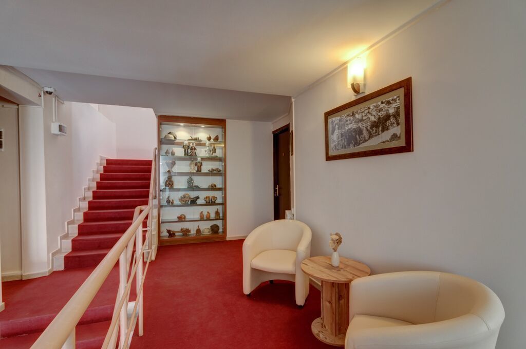 A staircase with red carpet leading to a room with a wooden floor, a glass display cabinet, two white armchairs, and a framed picture on the wall.