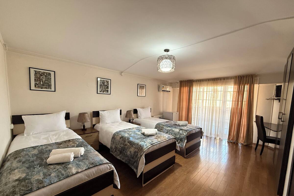 A hotel room with three beds, a desk, a chair, and a large window with curtains.