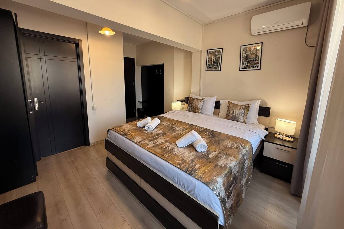A modern hotel room with a large bed, a desk, and a chair. There are two doors, one of which is open. The room has a ceiling light and an air conditioning unit.