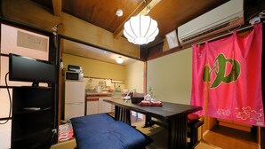 A traditional Japanese-style room with tatami flooring, a low table with a cushion, a television, and a kitchen area visible through an open door. - Oyado Sanjo Machiya (Kyoto)