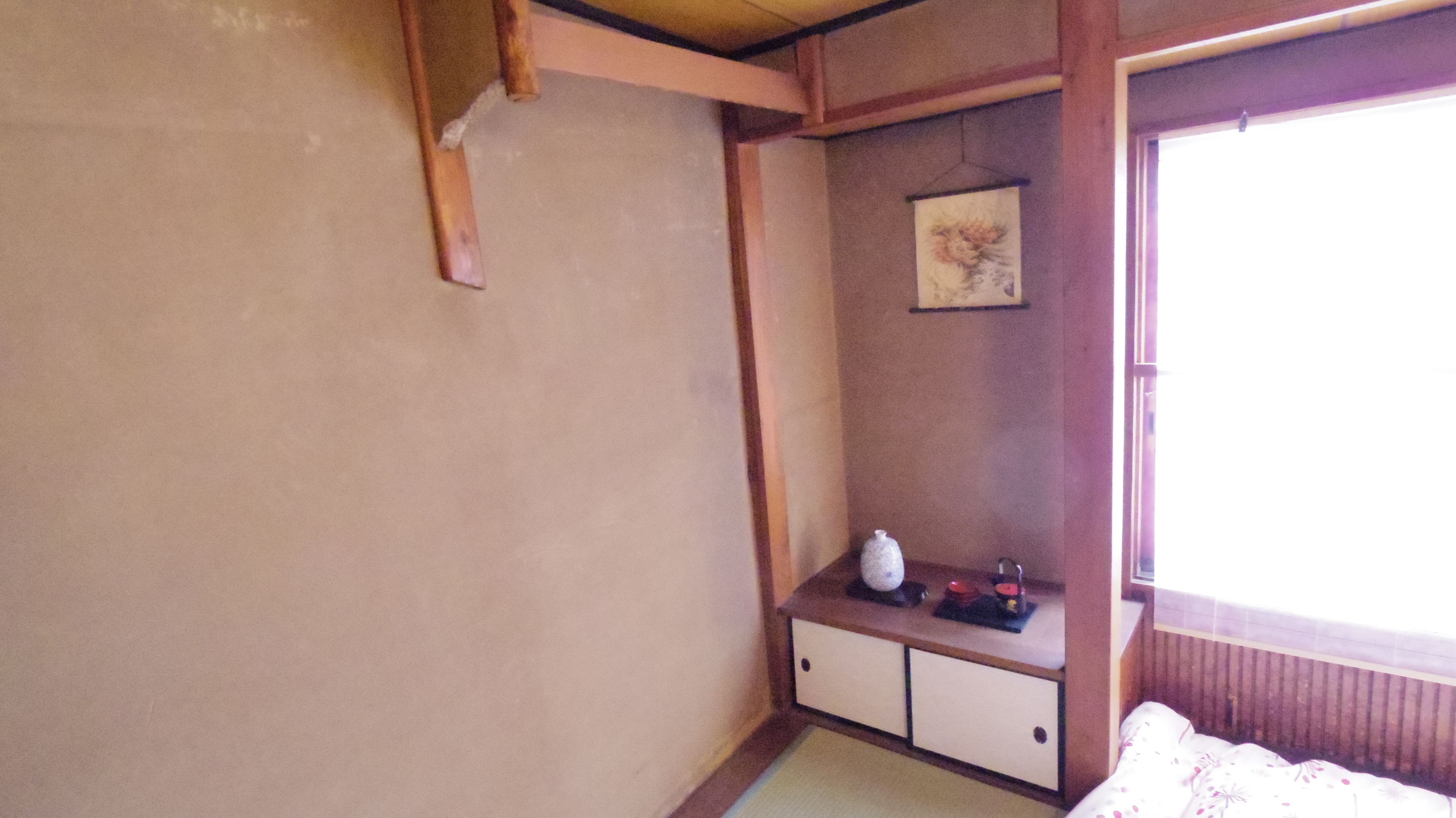 A room with a tatami mat floor, a low wooden cabinet with a small shelf, a framed picture on the wall, and a window with a view of the outside.