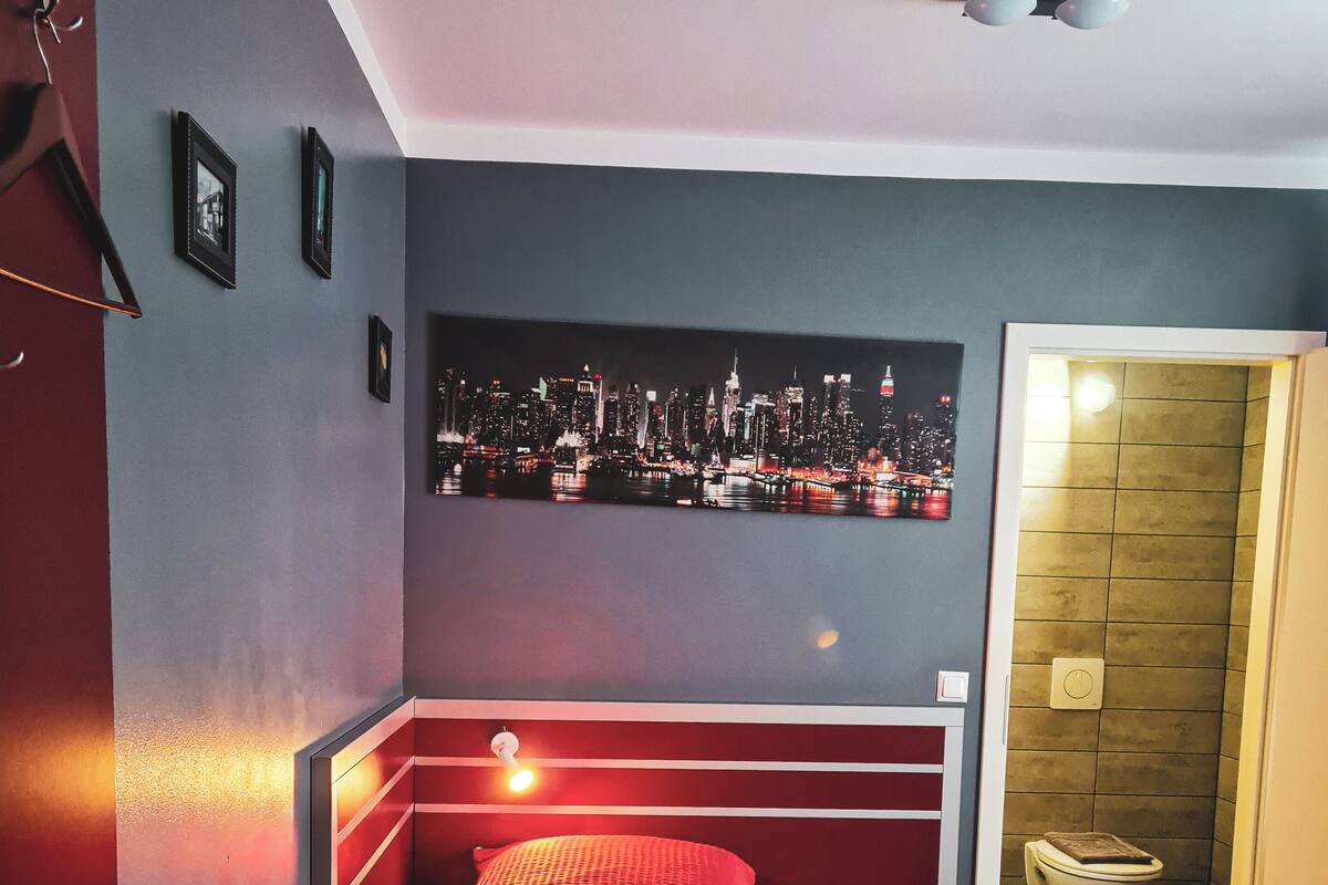 Quarto com cama, arte de parede com vista urbana, cabeceira vermelha e porta para o banheiro.