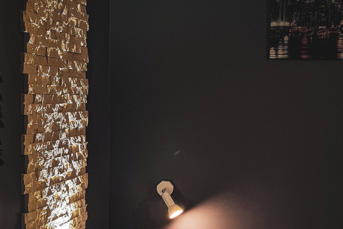 Um quarto com pouca luz, uma luminária na parede, uma cama com almofadas e um detalhe de parede com textura.