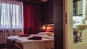 Een slaapkamer met een bed, een raam met gordijnen, en een badkamer met een wastafel en spiegel.