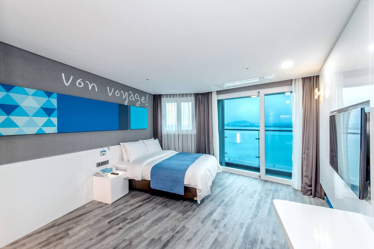 A modern hotel room with a large bed, a desk, and a view of the sea through a sliding door.