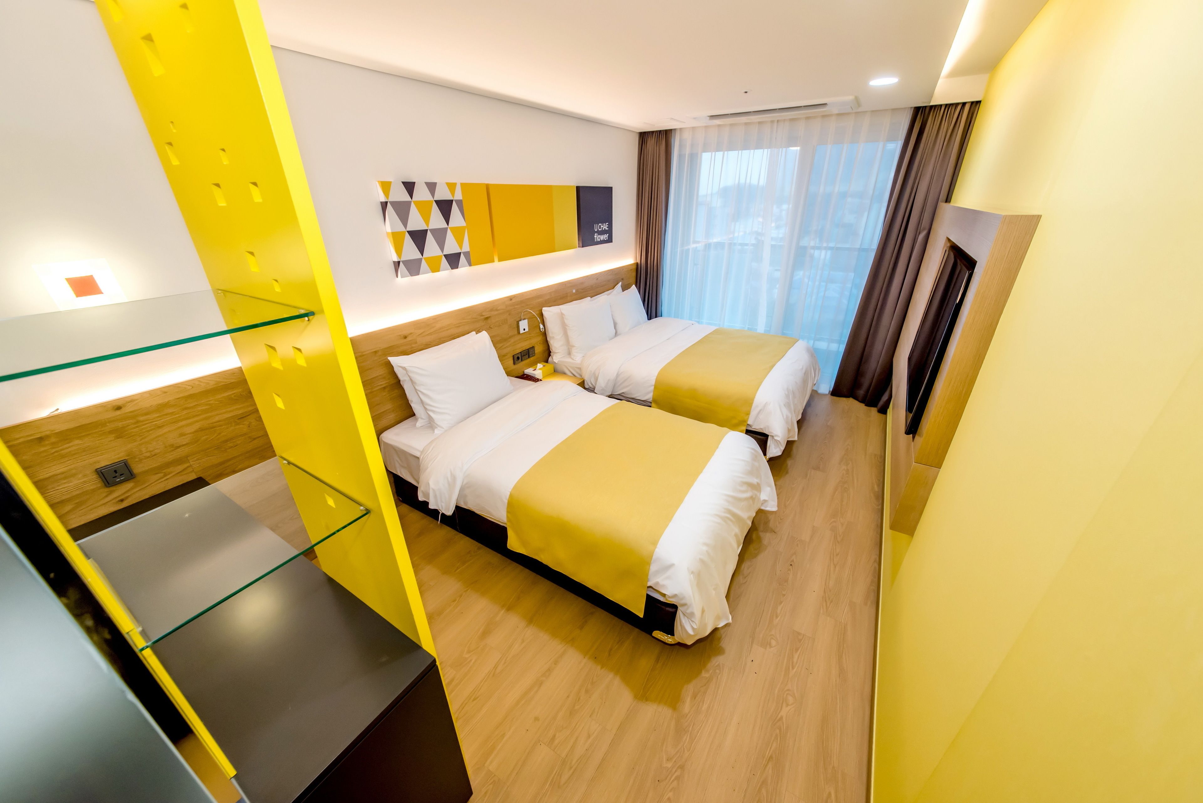 A hotel room with two beds, a glass partition, a wooden floor, and a modern decor with yellow accents.