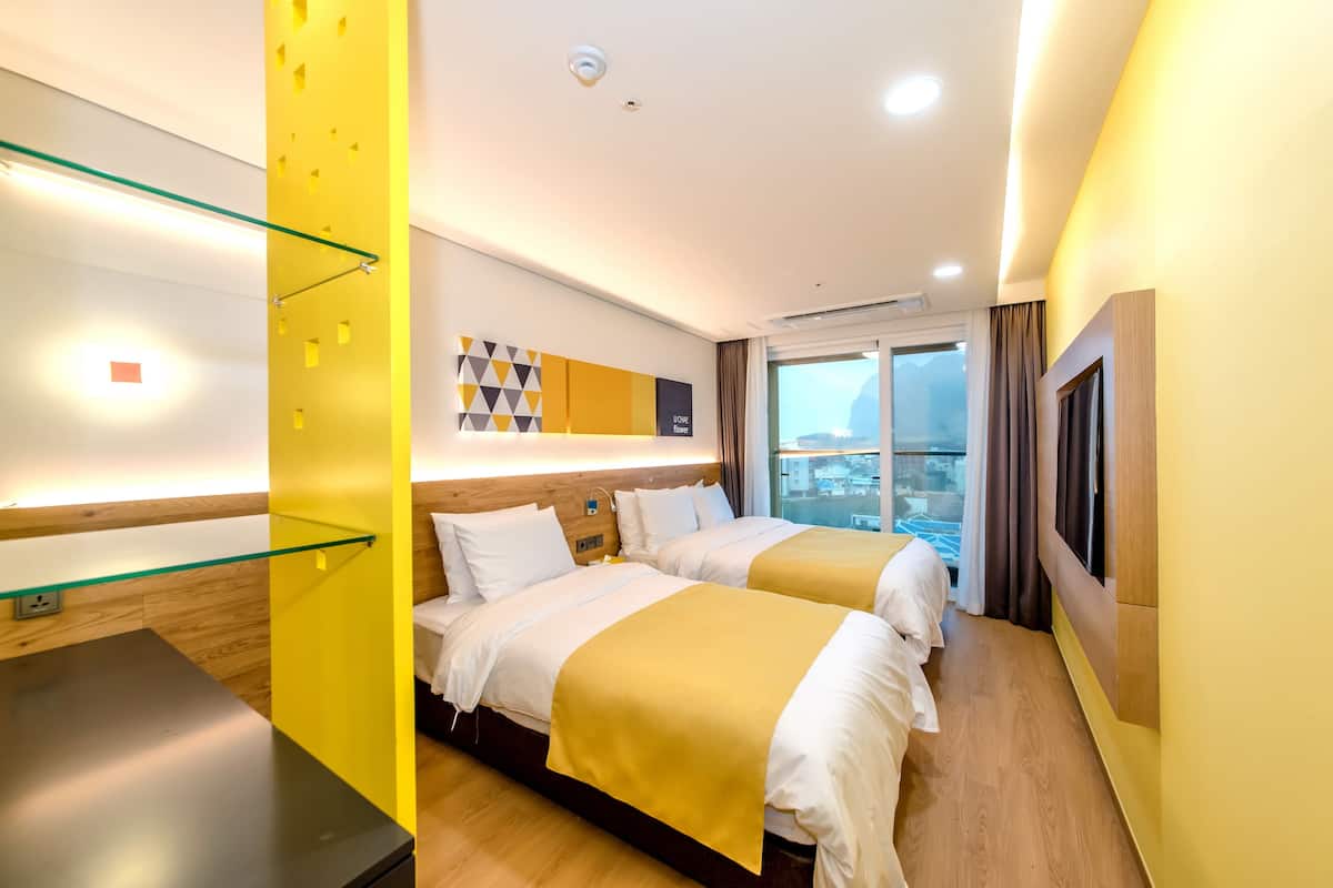 A hotel room with two beds, a glass partition, a wooden floor, a large window, and a TV mounted on the wall.