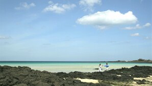 A beach with a rocky foreground and a person in a blue shirt and white pants.