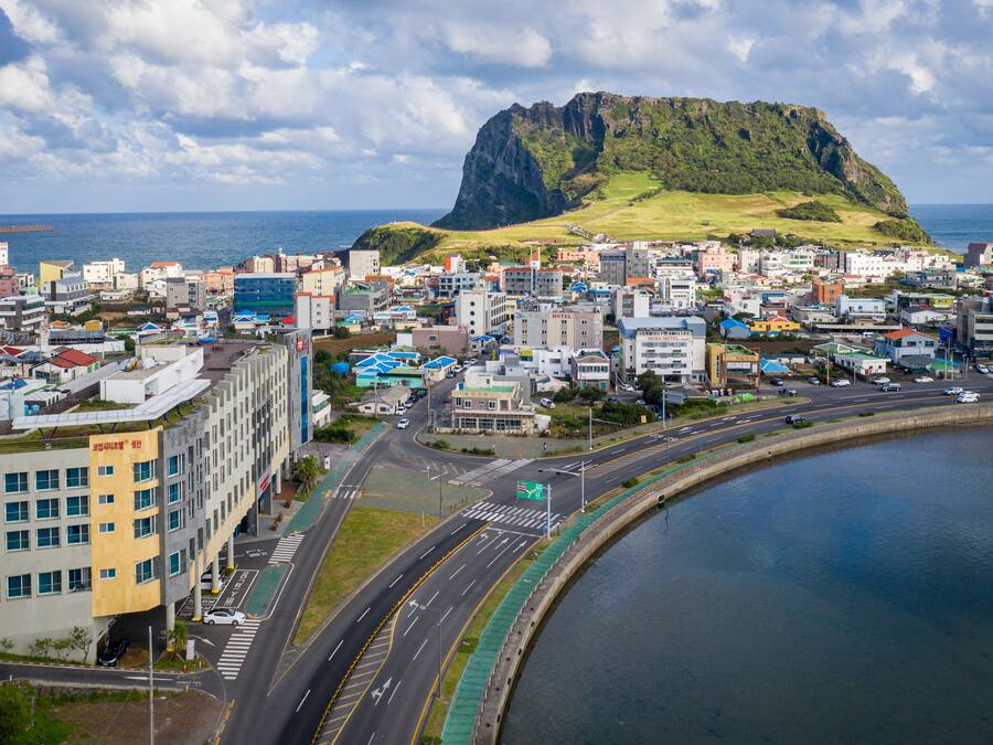 A coastal cityscape with a prominent mountain, modern buildings, and a curved road leading to a body of water.