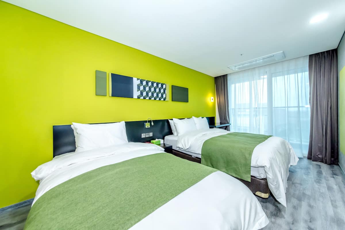 A hotel room with two beds, bright green walls, a modern headboard, and a large window with curtains.