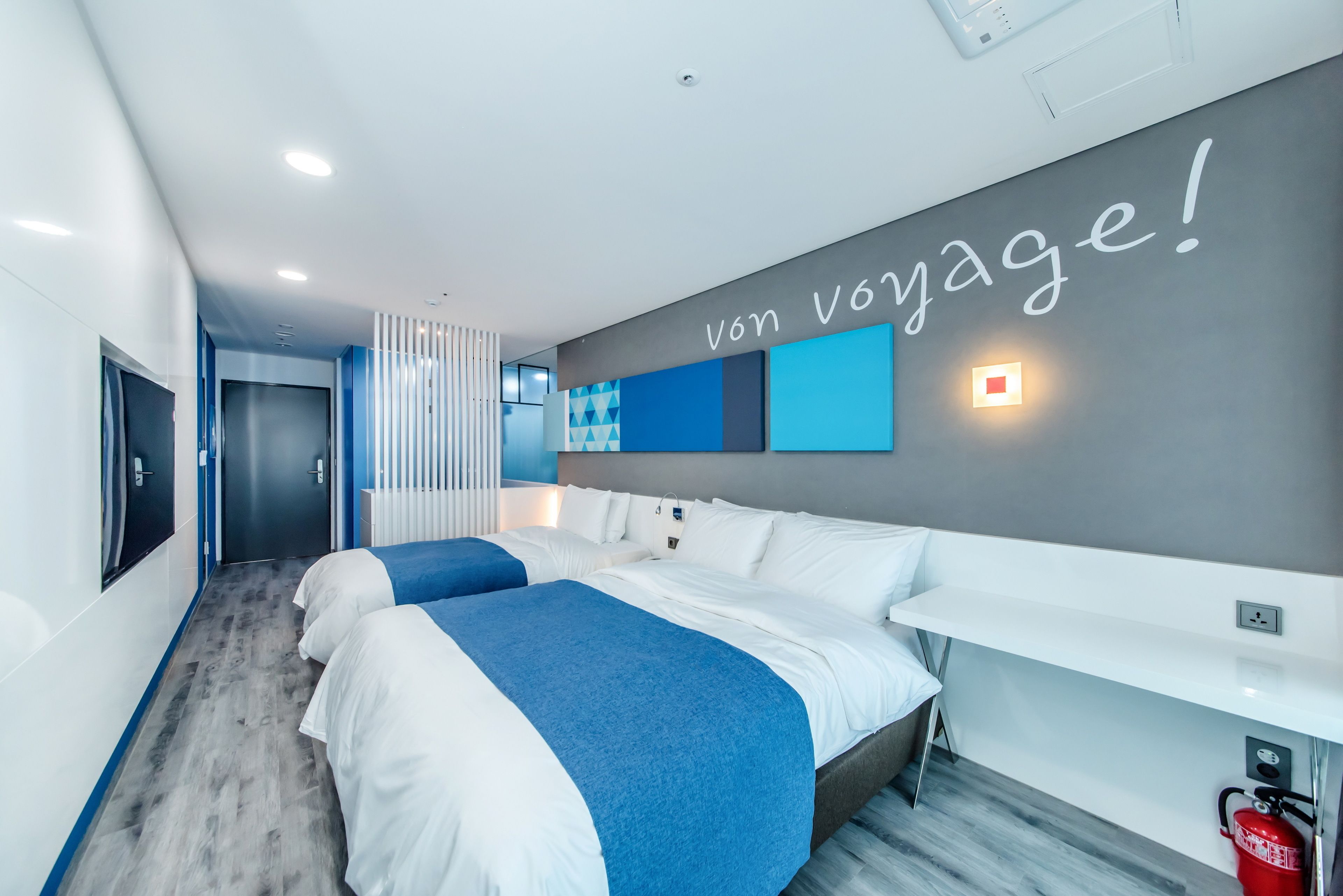 Ein modernes Hotelzimmer mit zwei Betten, einem großen Spiegel, einem Wandregal und einem Schild mit der Aufschrift 'Von Voyage!'.