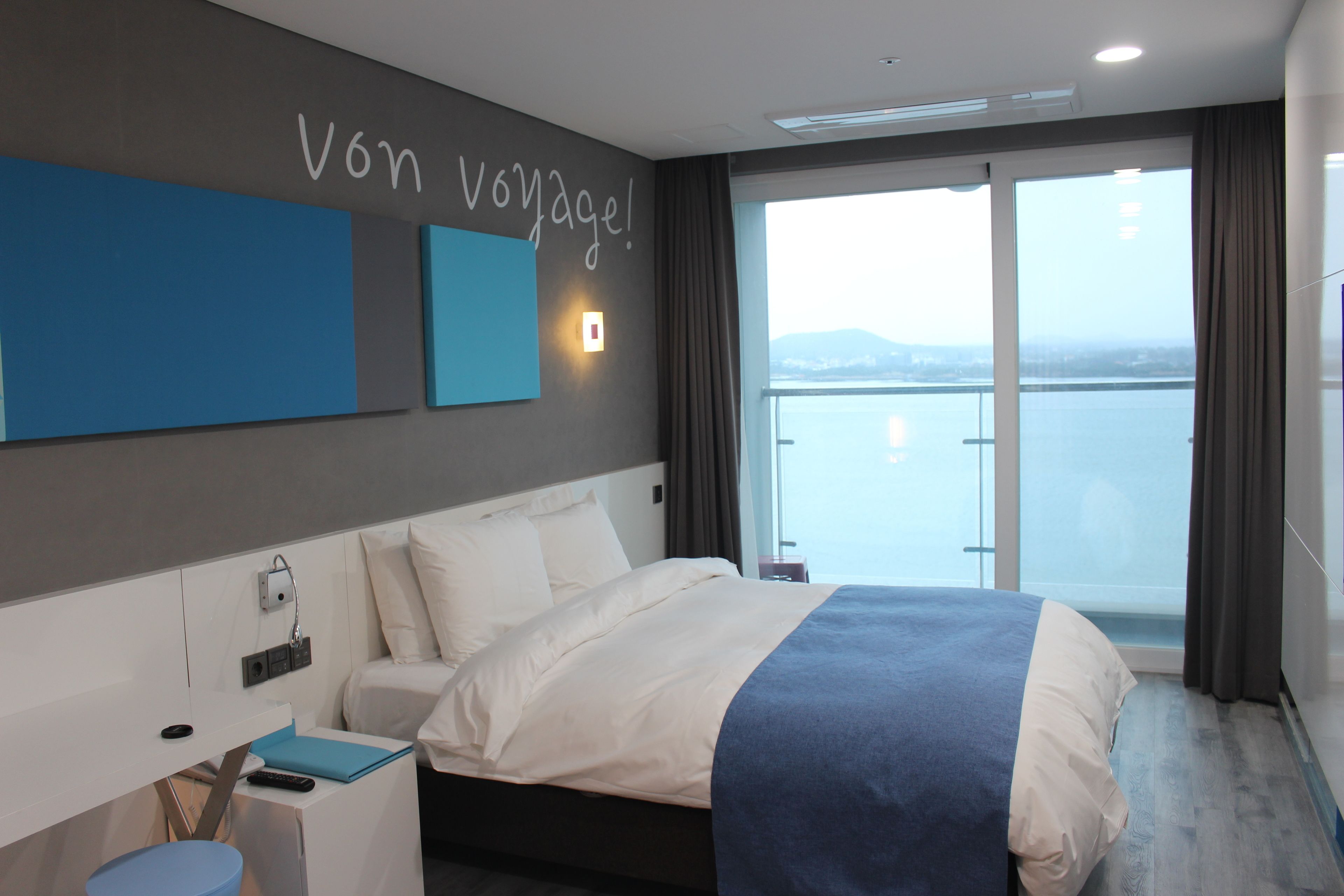 A modern hotel room with a large bed, a desk, and a view of the sea.