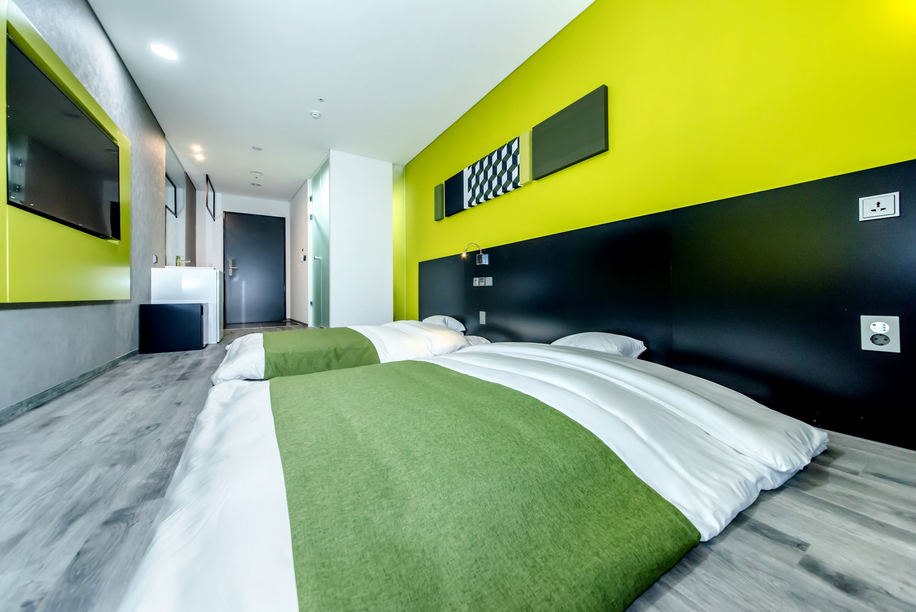 Habitació d'hotel moderna amb un llit gran, un edredó verd i blanc, un panell de capçalera negre i una paret d'accent groga brillant.