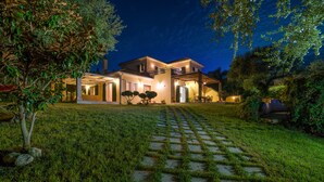 Casa de dos pisos con exterior bien iluminado, camino de entrada pavimentado y jardín con un árbol pequeño y arbustos.