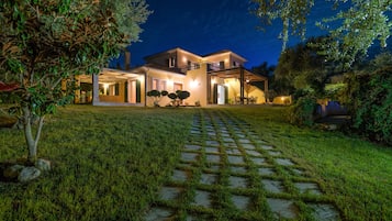 A two-story house with a well-lit exterior, a paved driveway, and a garden area with a small tree and shrubs.