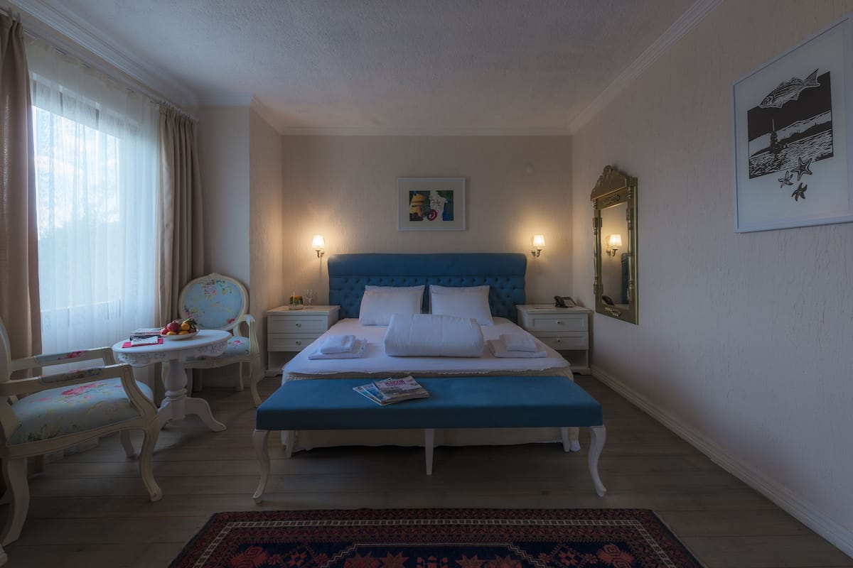 Un dormitorio con cabecera azul, ropa de cama blanca, una mesita de noche con lámpara, una silla y una pequeña mesa con frutero.