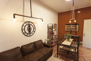 A living room with a brown leather sofa, a dining table with chairs, a metal chandelier, a wall clock, and a bookshelf. - Piazza Marina (Palermo)