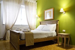Un lit bien fait, avec des draps blancs et une couverture à rayures, une table de chevet en bois avec une lampe, et un tableau encadré accroché au mur.