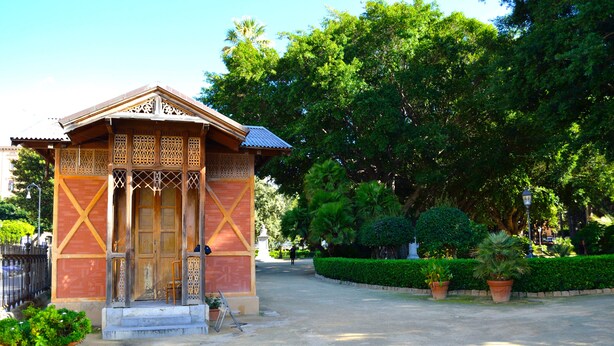 A small, traditional wooden building with intricate carvings, a tiled roof, and a paved entrance area with potted plants. - Piazza Marina (Palermo)