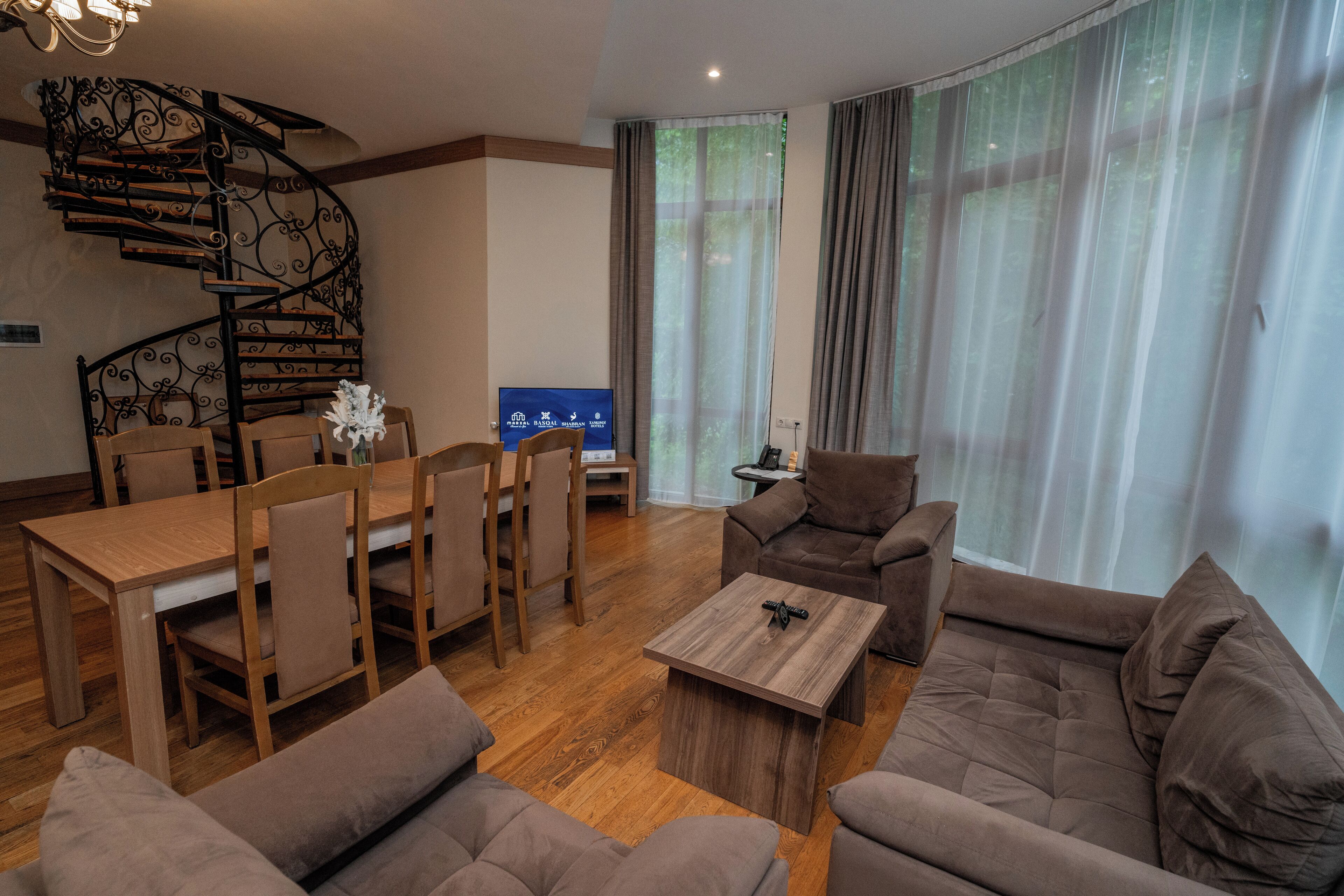 A modern living room with a wooden dining table, brown sofas, a coffee table, and a spiral staircase.