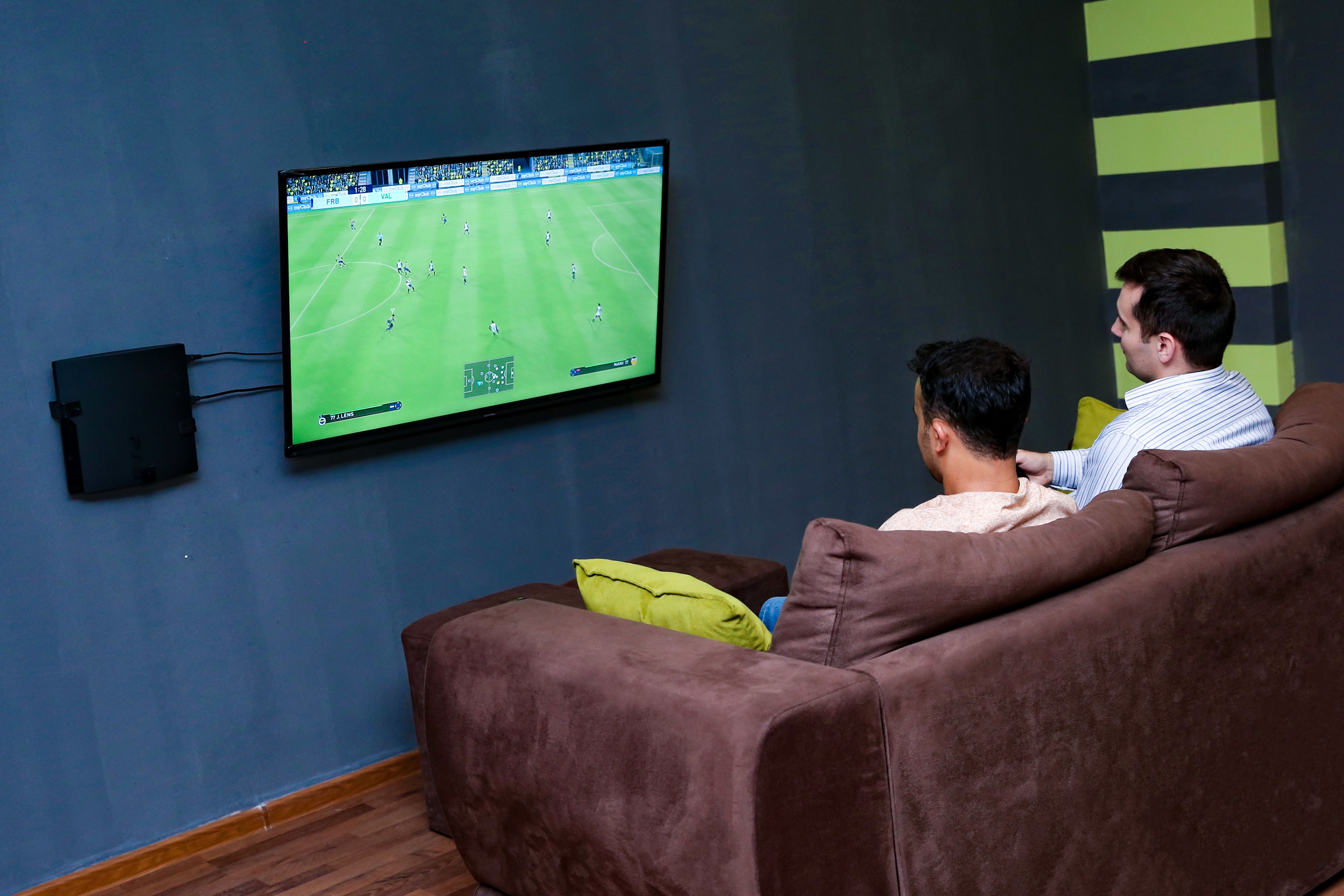 A room with a large flat-screen TV mounted on the wall displaying a soccer game. There is a brown sofa with a yellow pillow and a person sitting on it.