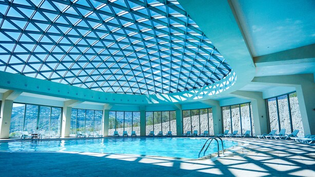 Ein Innenpool mit geometrischer Glasdecke, freiem Blick auf Berge und sonnendurchflutete Bereiche mit Liegestühlen.
