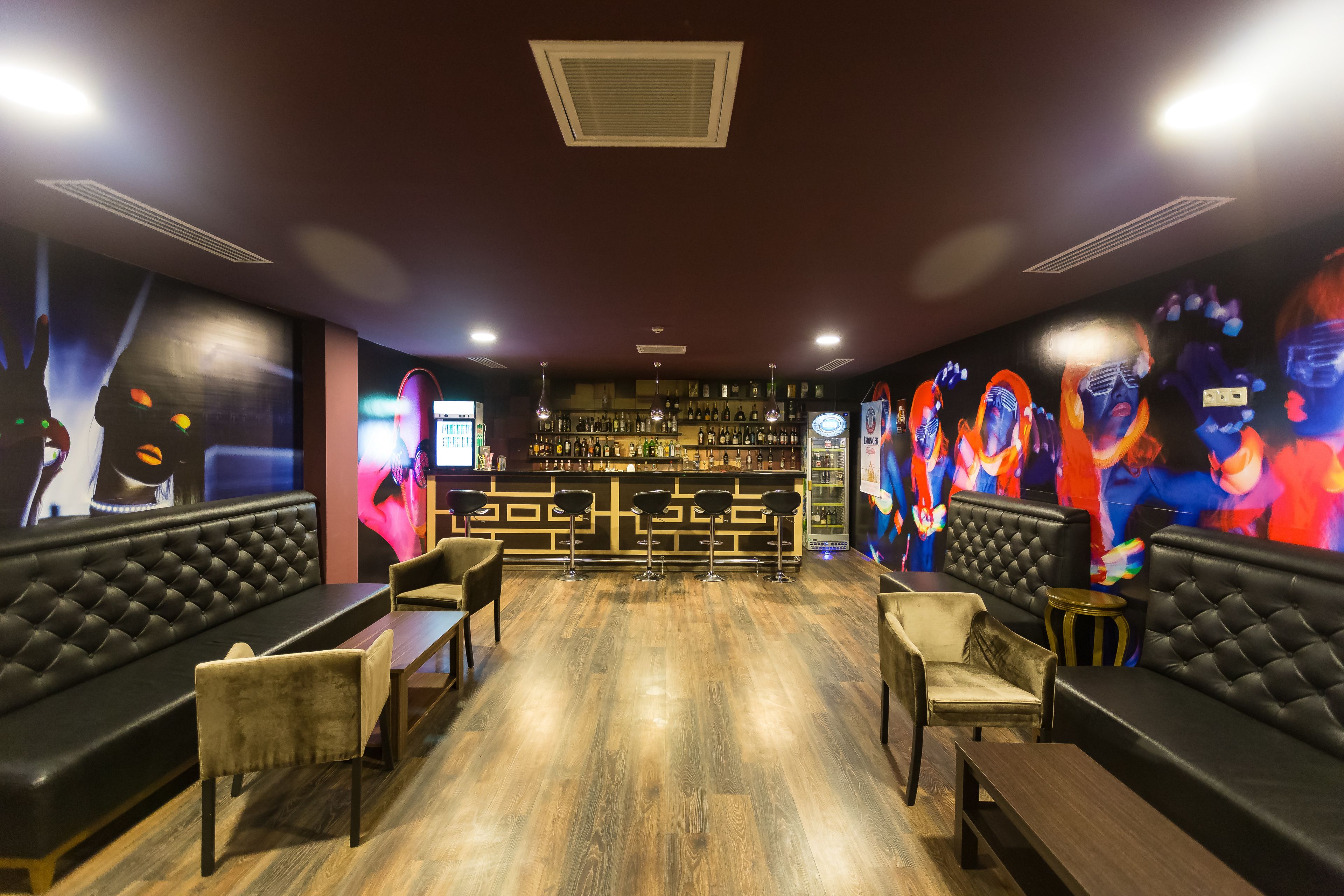 A bar area with a wooden floor, black leather sofas, and bar stools. There are shelves with bottles and a TV screen on the wall.