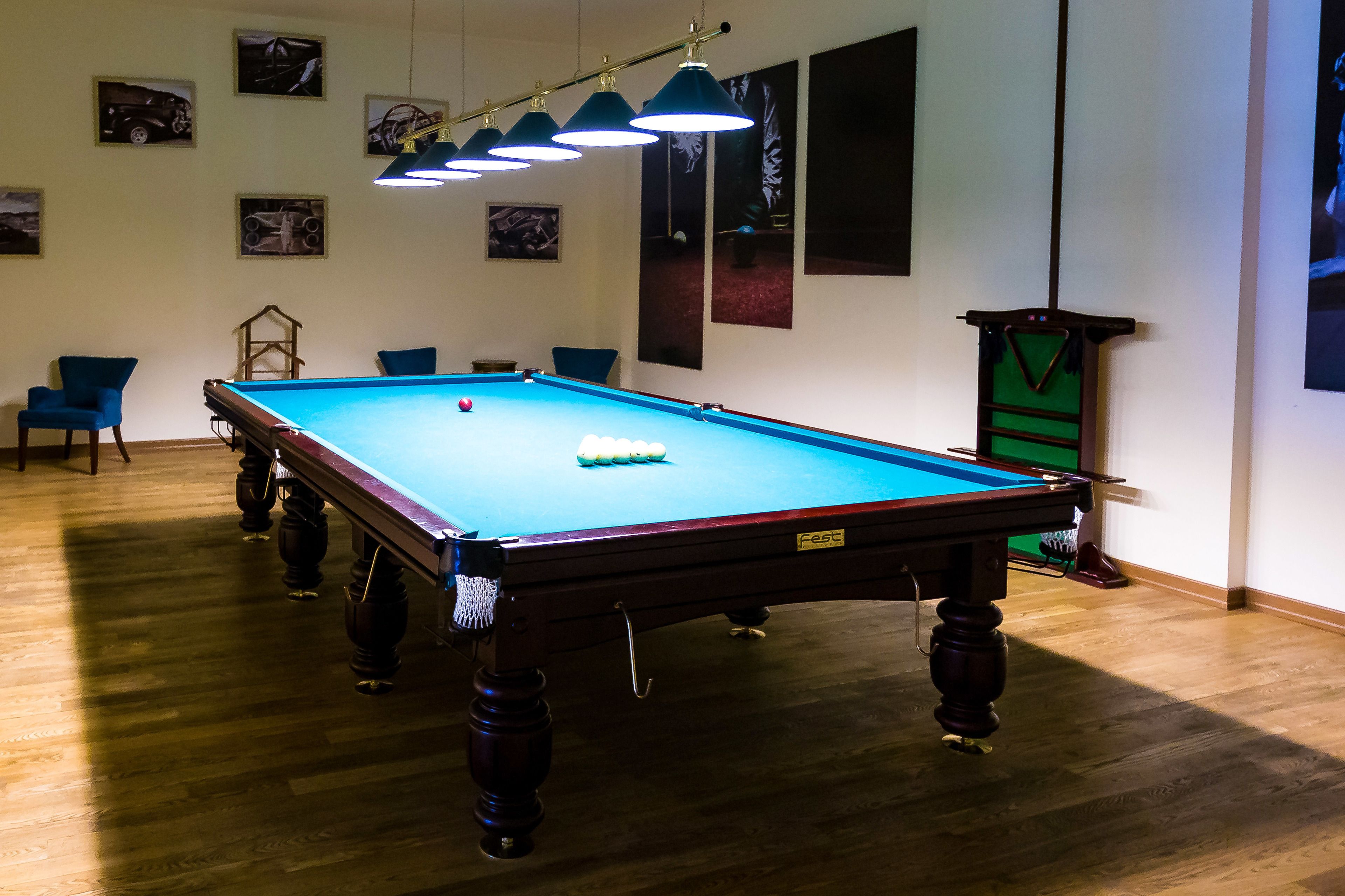 A pool table with a green felt surface, surrounded by blue chairs, in a room with wooden flooring and wall-mounted lights.