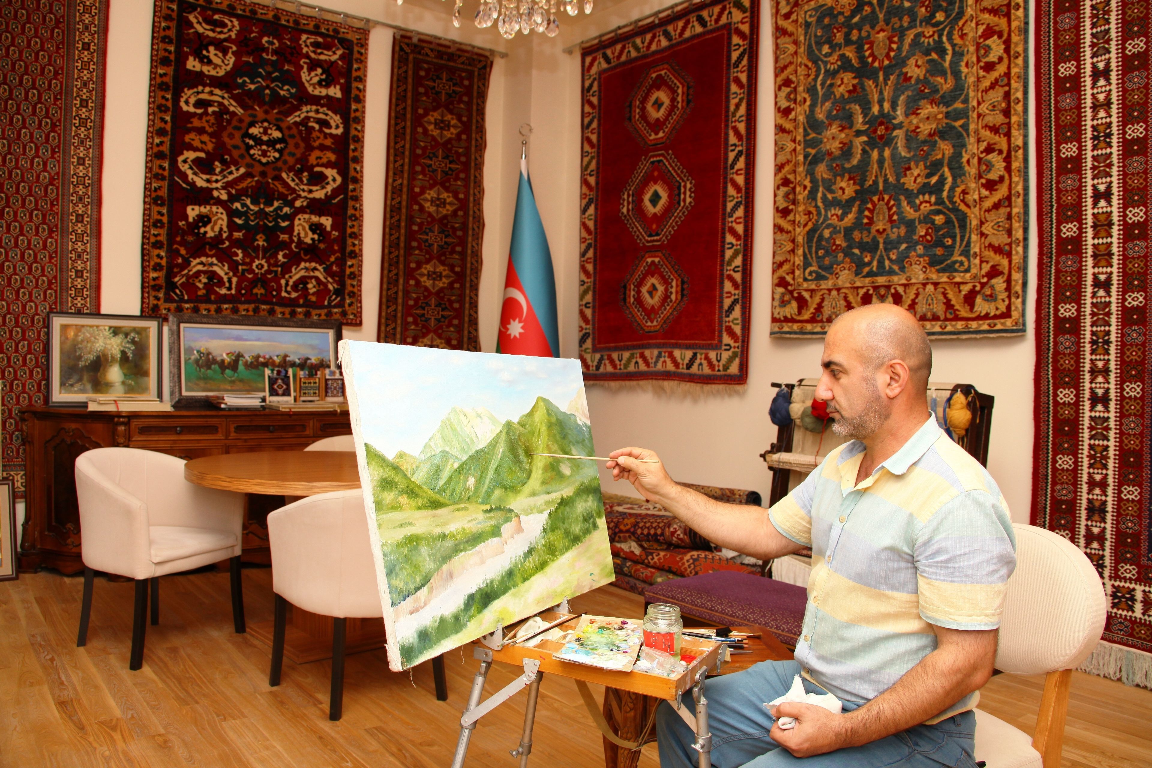 A man painting a landscape on a canvas in a room with traditional rugs and a flag.