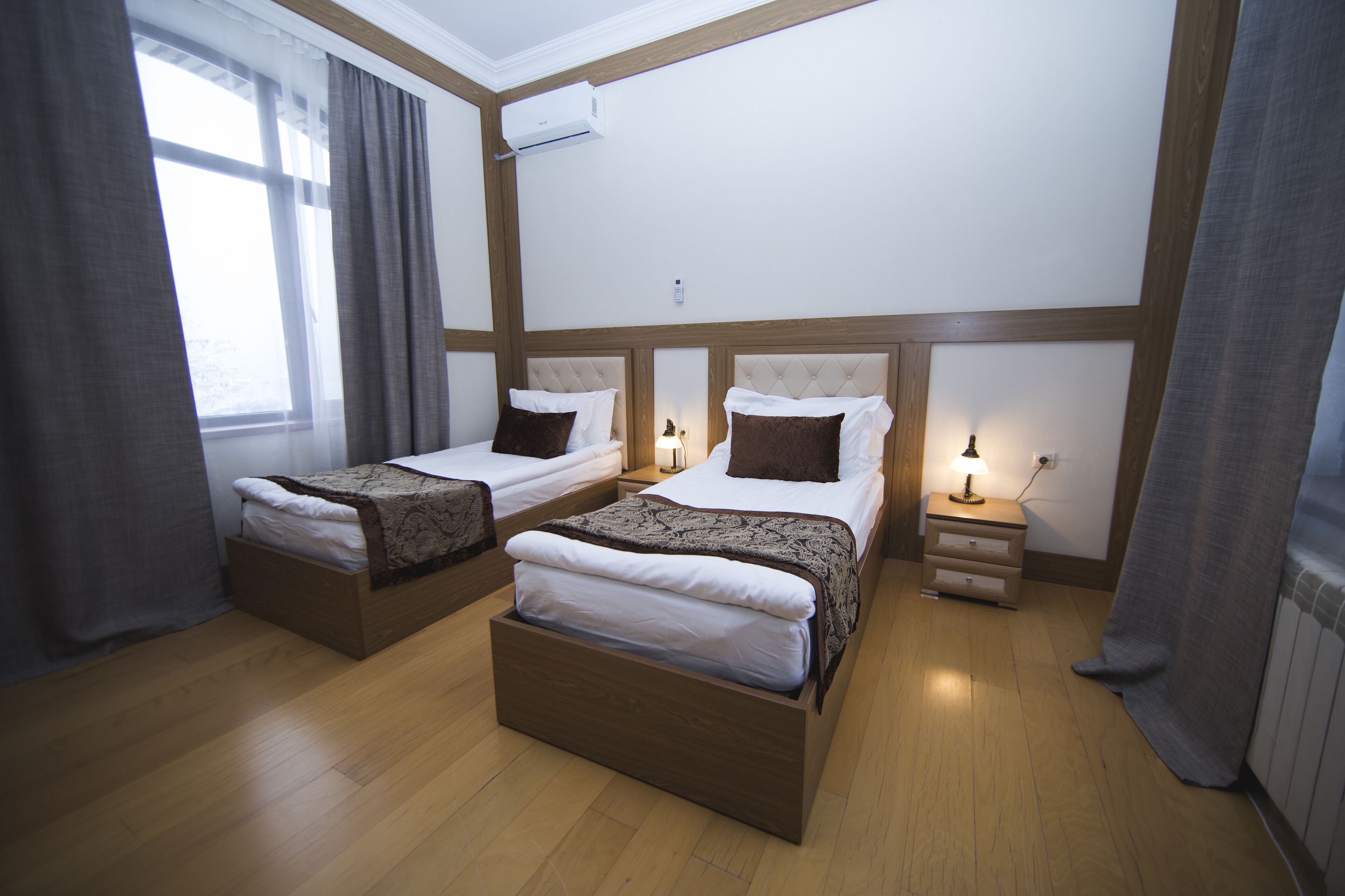 A hotel room with two beds, wooden headboards, bedside tables with lamps, and a window with curtains.