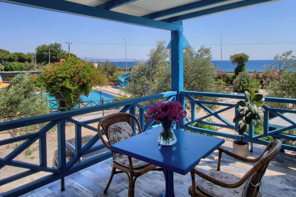 Un espacio exterior cubierto con mesa y sillas, vista al mar y una piscina al fondo.
