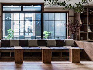 A modern interior with a long bench seating area, a wooden bookshelf, and a large window with a view of buildings. - Hotel Etwas Tenjin (Fukuoka)