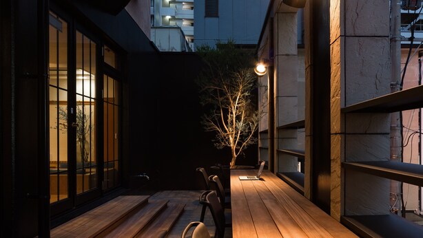An outdoor seating area with wooden benches and a laptop on a table, surrounded by a building with large windows and a tree. - Hotel Etwas Tenjin (Fukuoka)