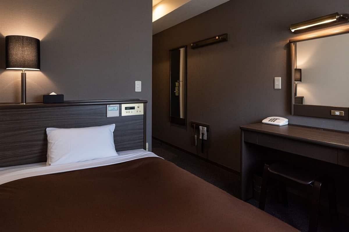 A hotel room with a bed, bedside lamp, desk, chair, and a mirror.