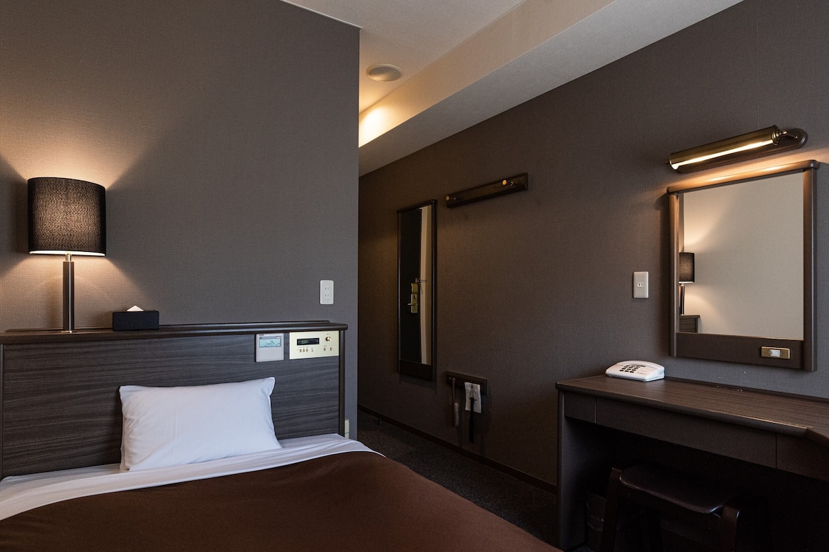 A hotel room with a bed, bedside lamp, control panel, desk, chair, and a mirror.