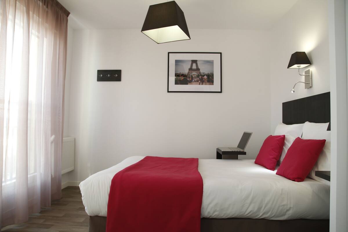 Een moderne hotelkamer met een groot bed, rood-witte beddengoed, een lijst met een afbeelding van de Eiffeltoren, een bureau met een laptop en een staande lamp.