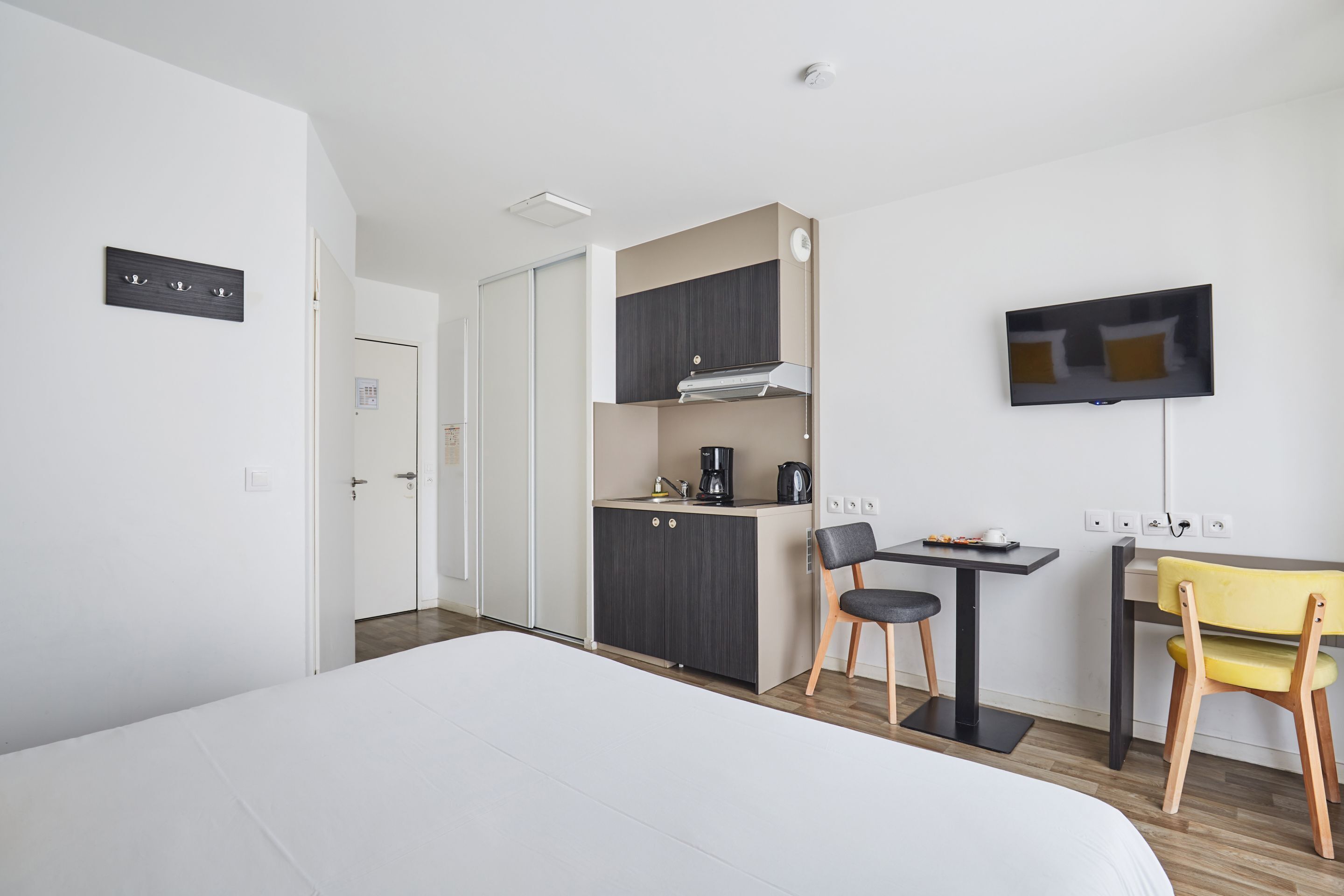 A modern hotel room with a large bed, a kitchenette, a dining area, and a TV mounted on the wall.