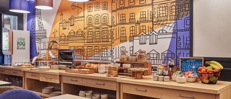 Un área de desayuno con mesas de madera, un mural de edificios y diferentes alimentos.