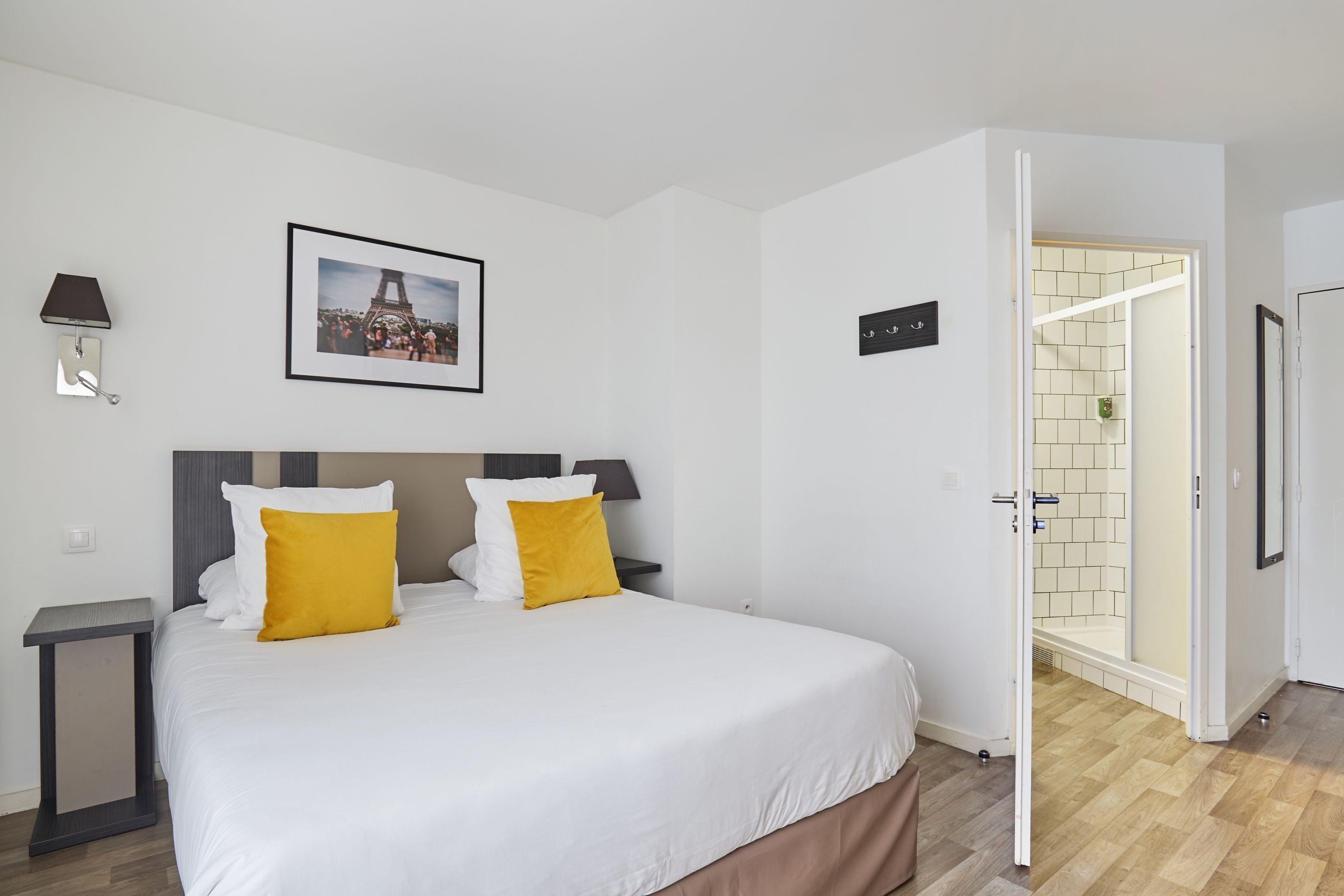 A hotel room with a bed, a nightstand, a picture of the Eiffel Tower, a bathroom with a shower, and a door leading to another room.