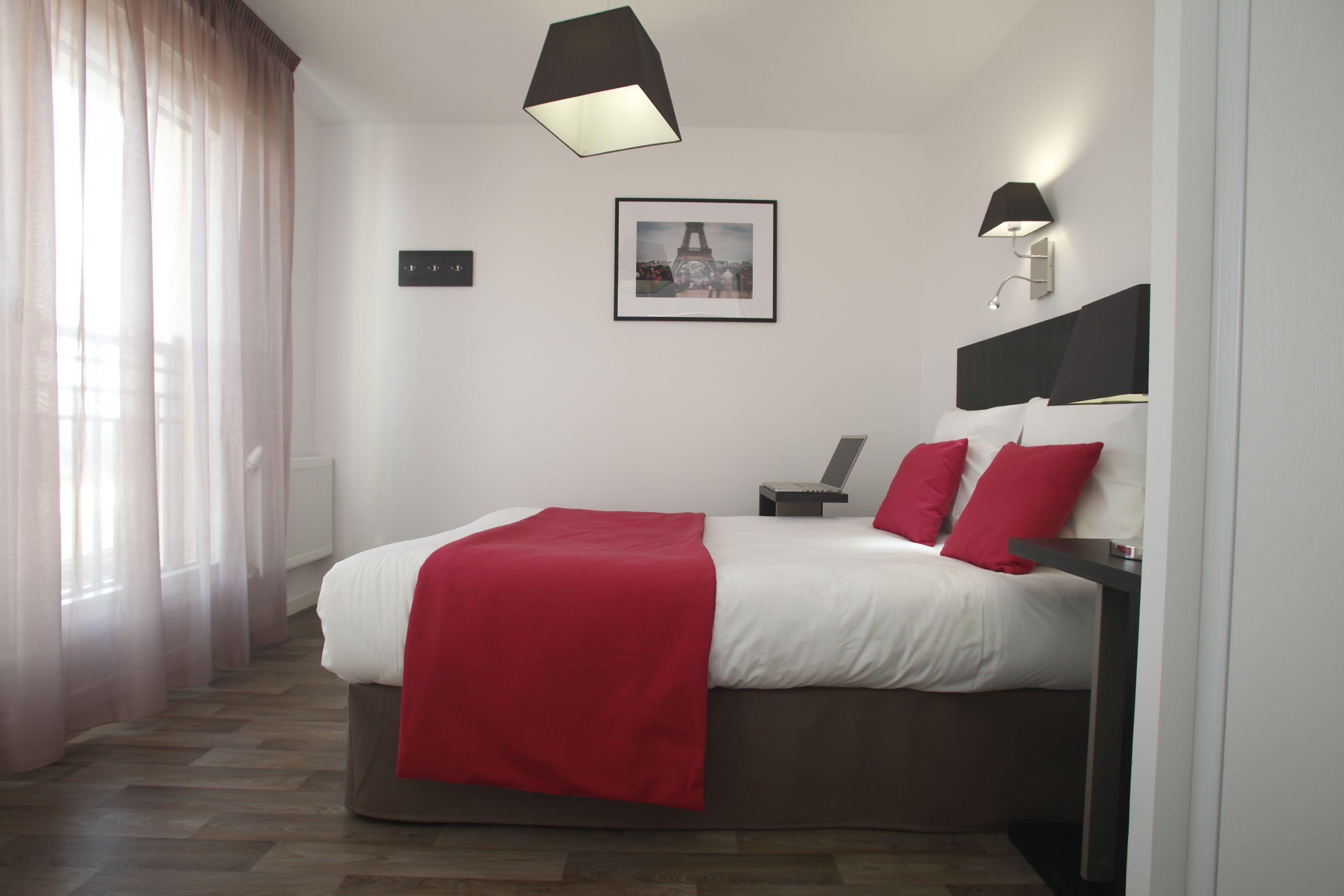 A modern hotel room with a large bed, a bedside table, a nightstand, a framed picture on the wall, and a window with sheer curtains.