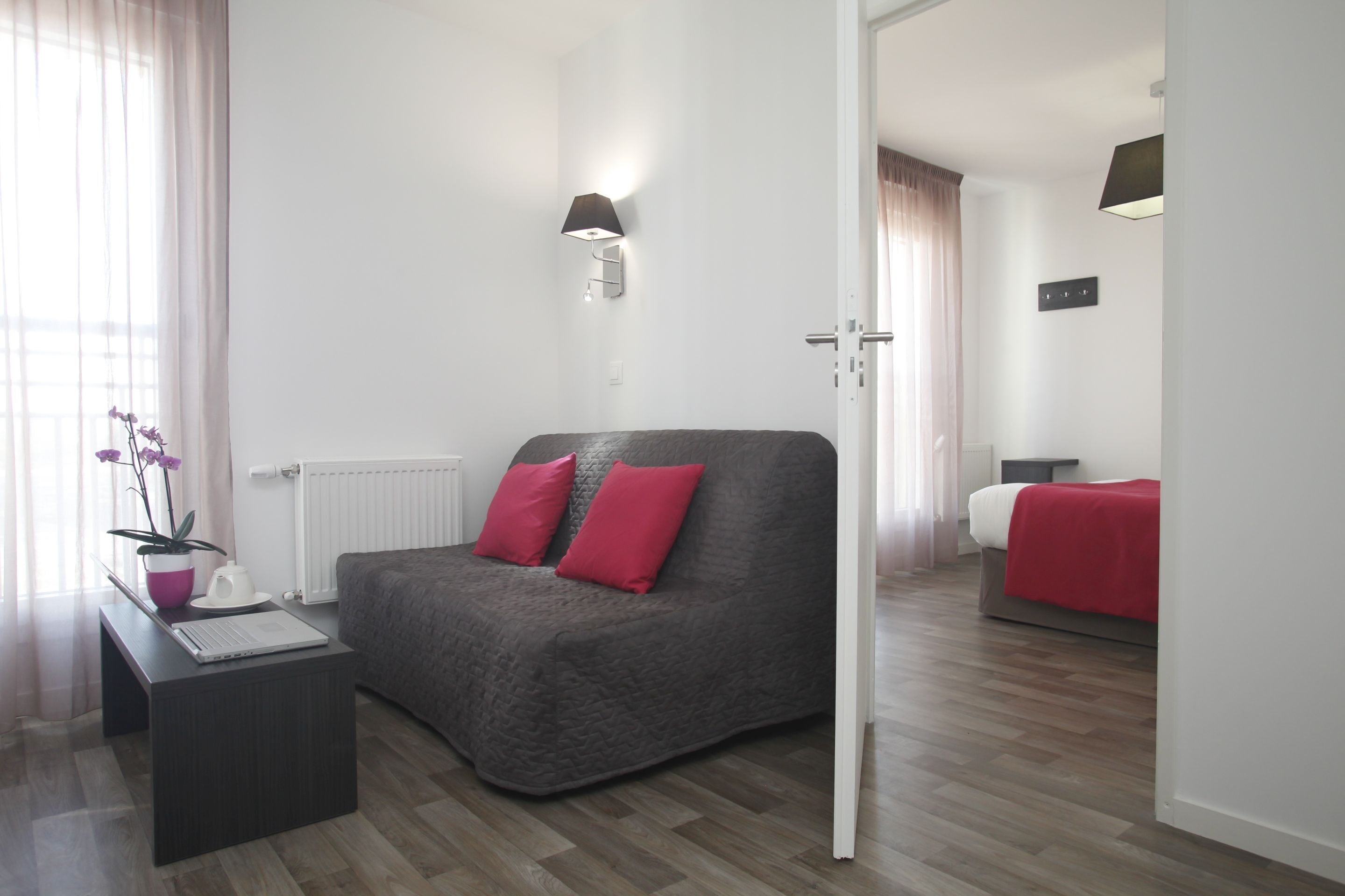 Un dormitorio moderno con un sofá gris, almohadas rojas, una mesita de noche con una computadora portátil y una planta en maceta.