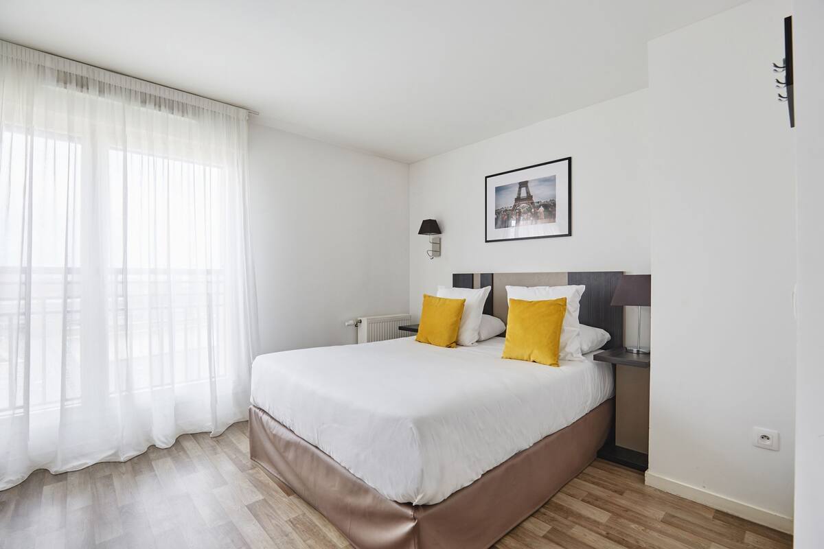 Een moderne hotelkamer met een groot bed, witte gordijnen, een ingelijst schilderij aan de muur en een nachtkastje met een lamp.